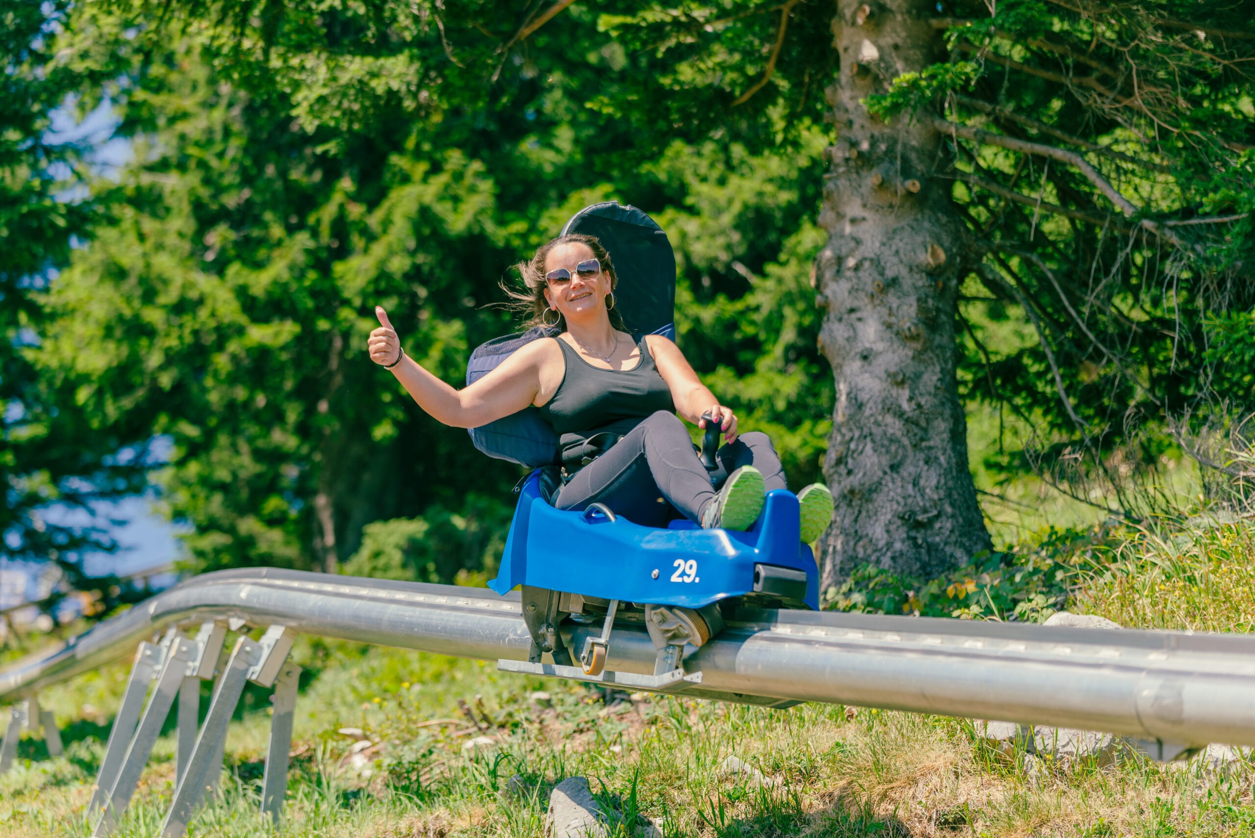 Woman with sunglasses rides a mountain roller coaster with a thumbs up. Warm summer day on the mountain